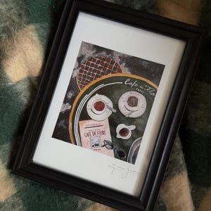 Puede incluir: Impresión artística enmarcada en negro que representa una escena de café con tazas de café, un menú y gafas de sol. La obra de arte se titula "Cafe de Flore, Paris" e incluye un diseño circular verde y rojo. El marco es negro.