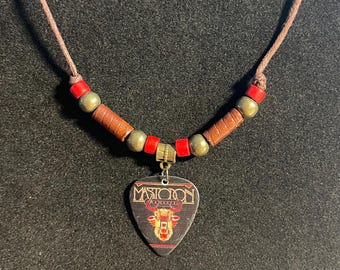 May include: A necklace featuring a guitar pick pendant with the text Mastodon and a graphic of a deer head. The cord is adorned with red cylindrical beads, metallic gold spherical beads, and brown cylindrical wooden beads.