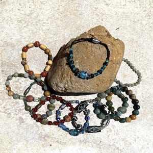 Op de afbeelding: Een collectie van elf kralenarmbanden gemaakt van diverse materialen, waaronder hout, glas en steen. De armbanden vertonen een scala aan kleuren zoals blauw, rood, groen, bruin en zwart, waarbij sommige zijn voorzien van metalen tussenstukjes.