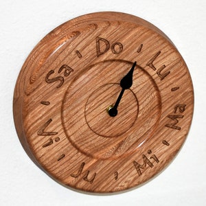 May include: A round wooden clock with a natural wood grain finish. The clock face features carved text and black hands. The text includes the words "Sa", "Do", "Lu", "Mi", "Ju", and "Vi".