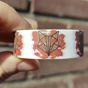 May include: A white washi tape with a repeating pattern of geometric fox heads in black outlines on a red watercolor background.