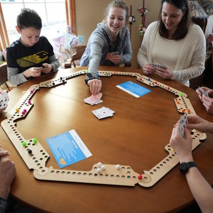 Peut inclure: Une famille joue à un jeu de société autour d'une grande table ronde en bois. Le jeu comprend une piste en bois avec des chevilles colorées et des billes. Les cartes sont distribuées et les joueurs sont engagés, créant une scène de plaisir et d'interaction familiale.