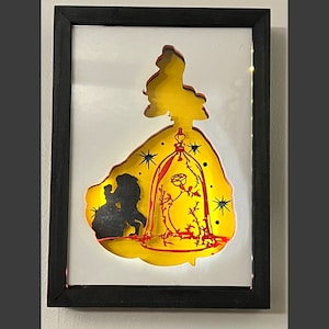 Pode incluir: Um recorte de papel amarelo de um vestido de mulher com uma silhueta de um casal dentro. O vestido é decorado com estrelas douradas e uma rosa vermelha dentro de uma cúpula de vidro.