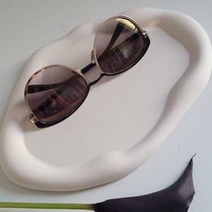 May include: A white ceramic tray with a unique, organic shape, featuring a pair of gold-rimmed sunglasses with brown lenses. A black calla lily with a green stem is in the foreground.