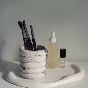 May include: A white marble-patterned ceramic makeup brush holder with four stacked rings, holding several black makeup brushes. The holder is on a white, wavy, ceramic tray with a clear glass bottle and a small, clear glass bottle with a black cap.