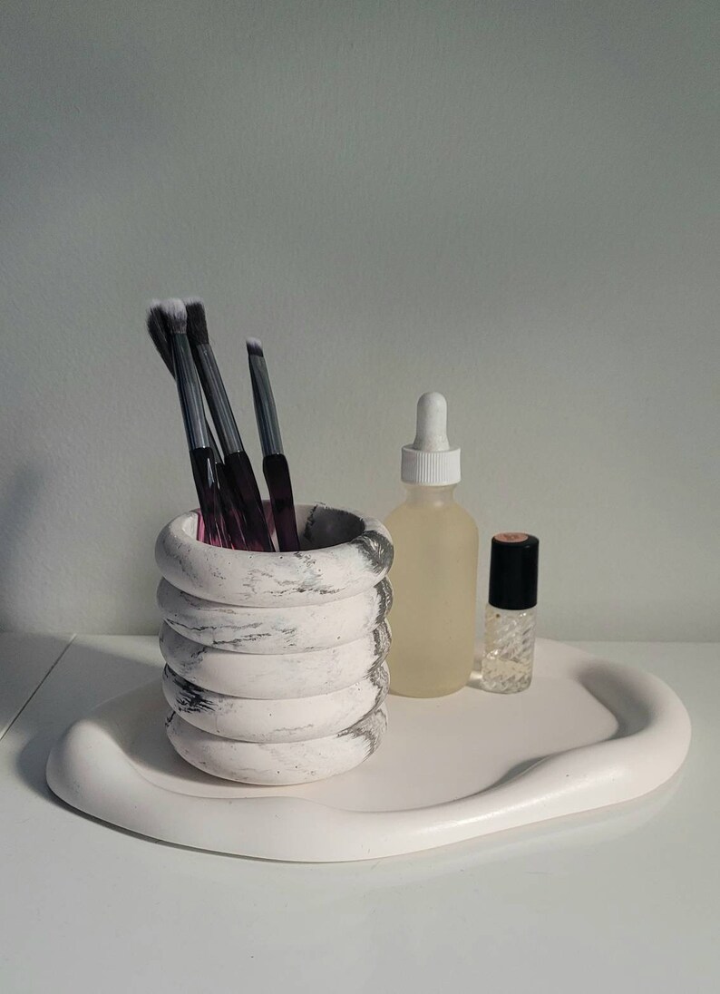 May include: A white marble-patterned ceramic makeup brush holder with four stacked rings, holding several black makeup brushes. The holder is on a white, wavy, ceramic tray with a clear glass bottle and a small, clear glass bottle with a black cap.