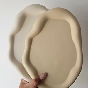 May include: Two ceramic trays in a light beige and cream color. The trays have an organic, wavy shape and are made of a smooth, matte material.