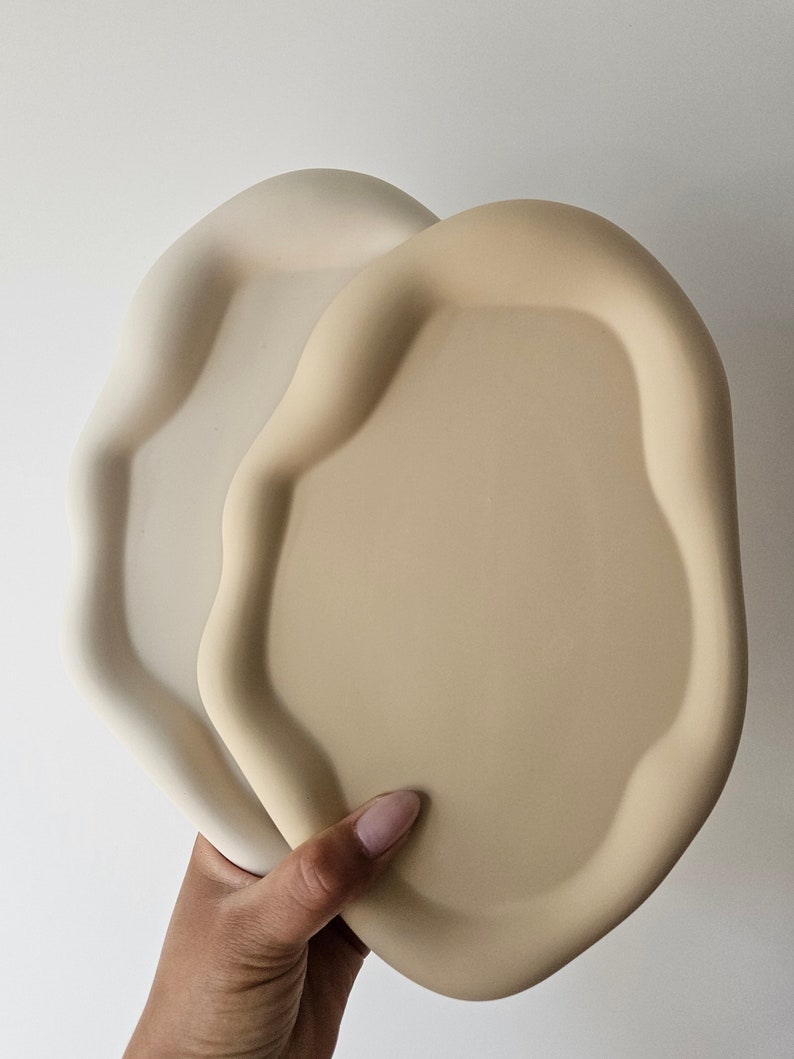 May include: Two ceramic trays in a light beige and cream color. The trays have an organic, wavy shape and are made of a smooth, matte material.
