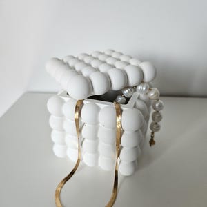 May include: A white ceramic jewellery box with a textured, bubble-like design. The box is open and a gold chain necklace and a pearl necklace are inside.
