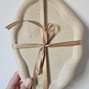 May include: A beige ceramic tray with an irregular shape, tied with a brown ribbon. The tray has a speckled texture.