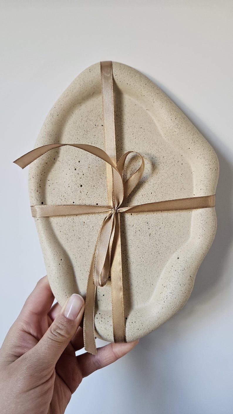 May include: A beige ceramic tray with an irregular shape, tied with a brown ribbon. The tray has a speckled texture.