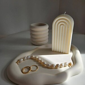 May include: A white ceramic tray with a wavy edge, featuring a white candle with a geometric design, a small white ceramic container, a pearl necklace, and two gold rings.