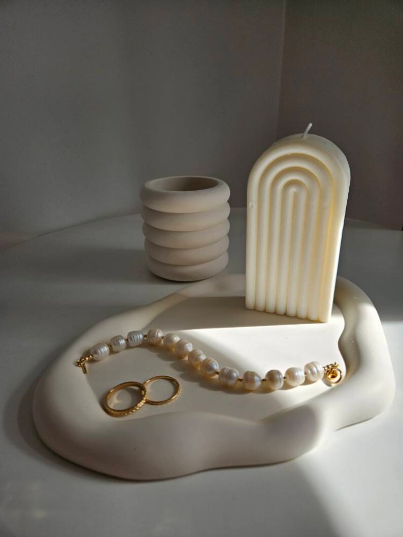 May include: A white ceramic tray with a wavy edge, featuring a white candle with a geometric design, a small white ceramic container, a pearl necklace, and two gold rings.
