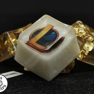 May include: A white resin keycap with a gold and blue League of Legends logo. The keycap is surrounded by smaller gold resin keycaps.