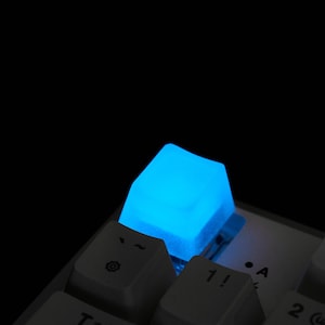May include: Close-up of a glowing blue keyboard key. The key is illuminated against a dark background, with other white keys visible. The key is a light blue color and appears to be made of translucent material.