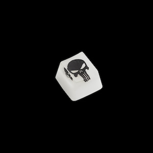 May include: A white keycap with a black skull graphic. The skull design is centered on the top surface of the keycap. The keycap is square and appears to be made of plastic.