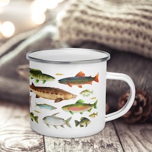Può includere: Tazza in smalto bianco con bordo argentato, decorata con illustrazioni ad acquerello di vari pesci. I pesci sono in tonalità di verde, marrone, blu e arancione. La tazza ha un manico bianco ed è appoggiata su una superficie di legno.