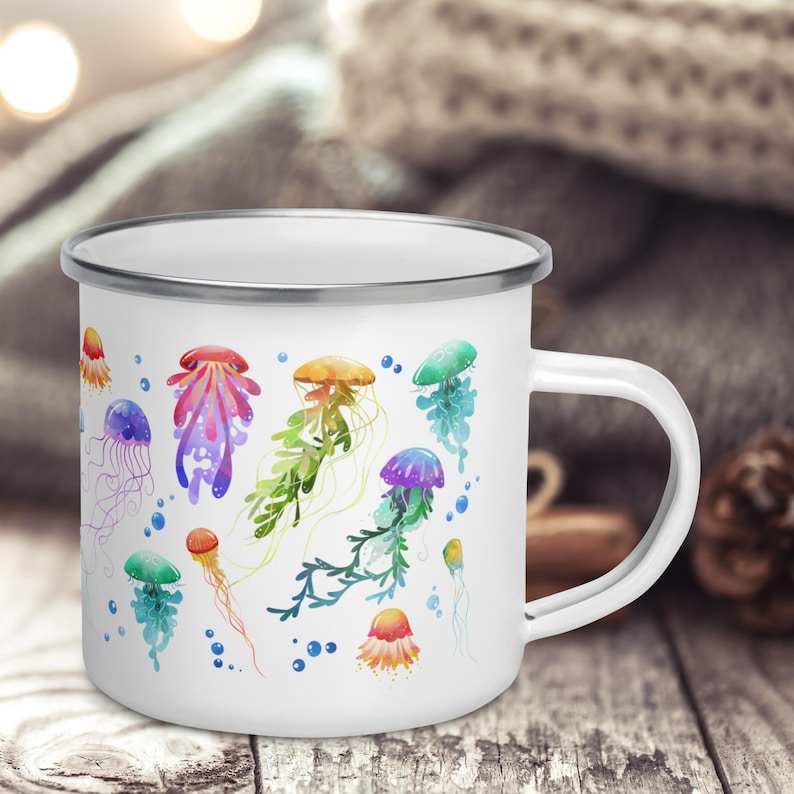 Pu&ograve; includere: Tazza in smalto bianco con un design ad acquerello colorato di meduse e alghe. La tazza ha un bordo e un manico argentati.