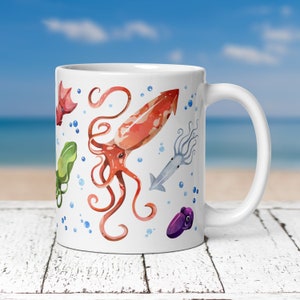 May include: White ceramic mug featuring watercolor illustrations of various sea creatures, including a red squid, a green squid, and a purple cuttlefish. Blue bubbles are scattered around the sea creatures. The mug has a white handle.