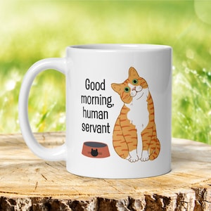 A white ceramic mug depicting an orange tabby cat waiting expectantly by its empty food bowl, with the text "Good morning, human servant" printed above the bowl.