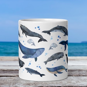 11-ounce ceramic mug with a watercolour whale print rests on a table with the ocean behind it. The centre of the mug is displayed. Visible on this side of the mug are a blue whale, minke whale, two beluga whales, a humpback whale, and a sperm whale.