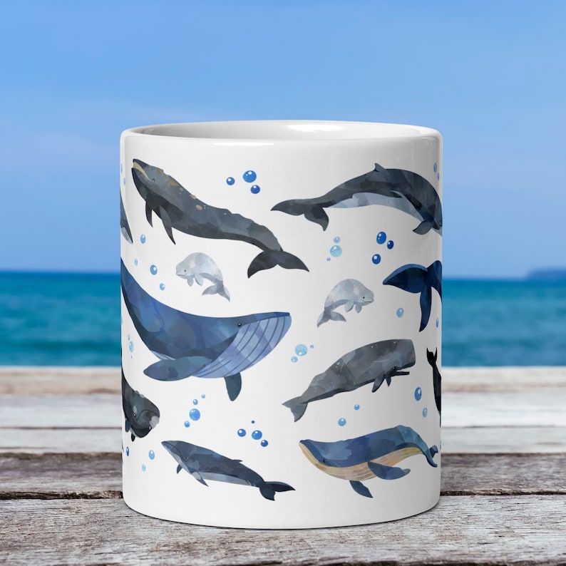 11-ounce ceramic mug with a watercolour whale print rests on a table with the ocean behind it. The centre of the mug is displayed. Visible on this side of the mug are a blue whale, minke whale, two beluga whales, a humpback whale, and a sperm whale.