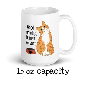 May include: White ceramic mug with a cartoon orange tabby cat and the text "Good morning, human servant". The cat is looking at the viewer and has a bowl of cat food in front of it. The mug has a 15 oz capacity.