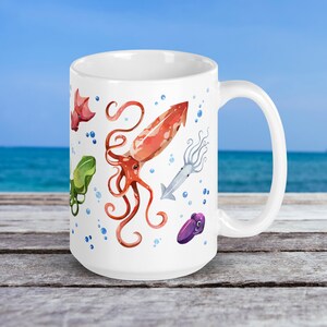 May include: White ceramic mug featuring watercolor illustrations of various sea creatures, including a red squid, a green octopus, and a purple cuttlefish. Blue bubbles accent the design. The mug is set against a backdrop of the ocean.