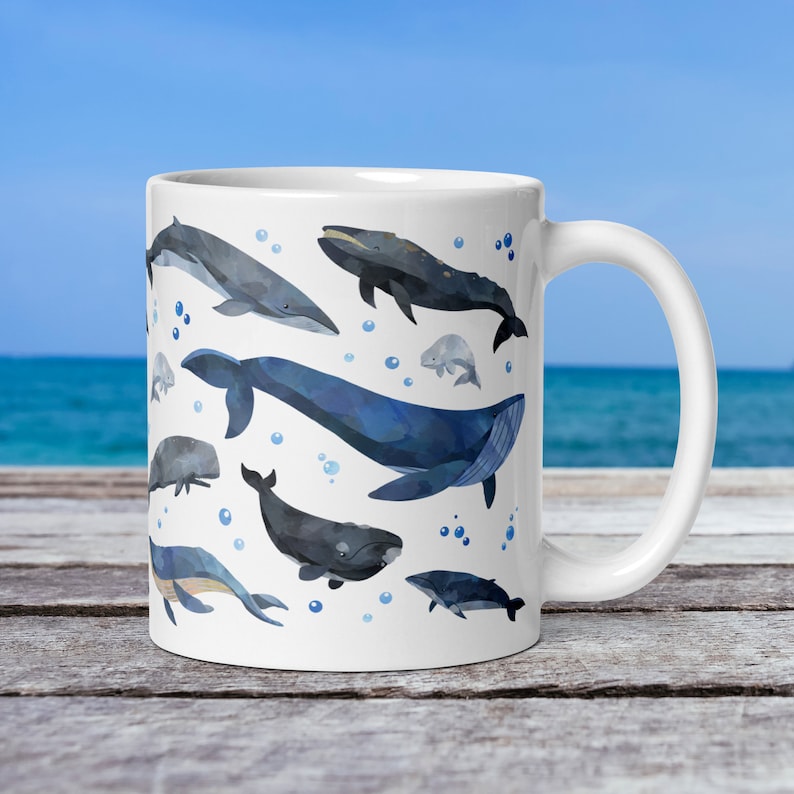 11-ounce ceramic mug with a watercolour whale print rests on a table with the ocean behind it. The mug's handle is on the right. A blue whale is at the center of this side, with a right whale, fin whale, grey whale, and minke whale also visible.