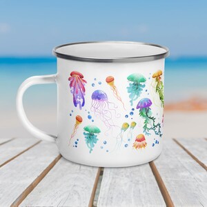A 12-ounce enamel mug rests on an outdoor table with the ocean behind it. The mug has a silver rim and its handle is on the left. Printed on the white mug are colourful watercolour illustrations of jellyfish in pink, green, orange, purple and blue.