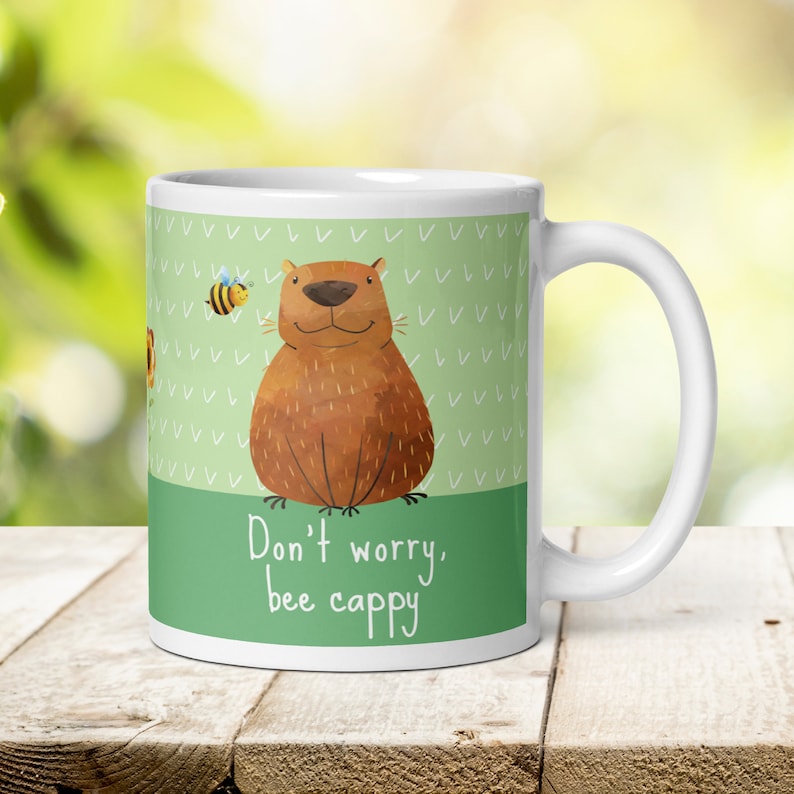 Op de afbeelding: Witte keramische mok met een groene achtergrond en een cartoonillustratie van een bruine beer met een bij die ernaast vliegt. De tekst "Don't worry, bee cappy" staat op de mok gedrukt.