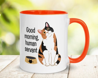 May include: White ceramic mug with an orange rim and handle. The mug features a cartoon calico cat sitting with the text "Good morning, human servant" and a cat food bowl with a cat silhouette.
