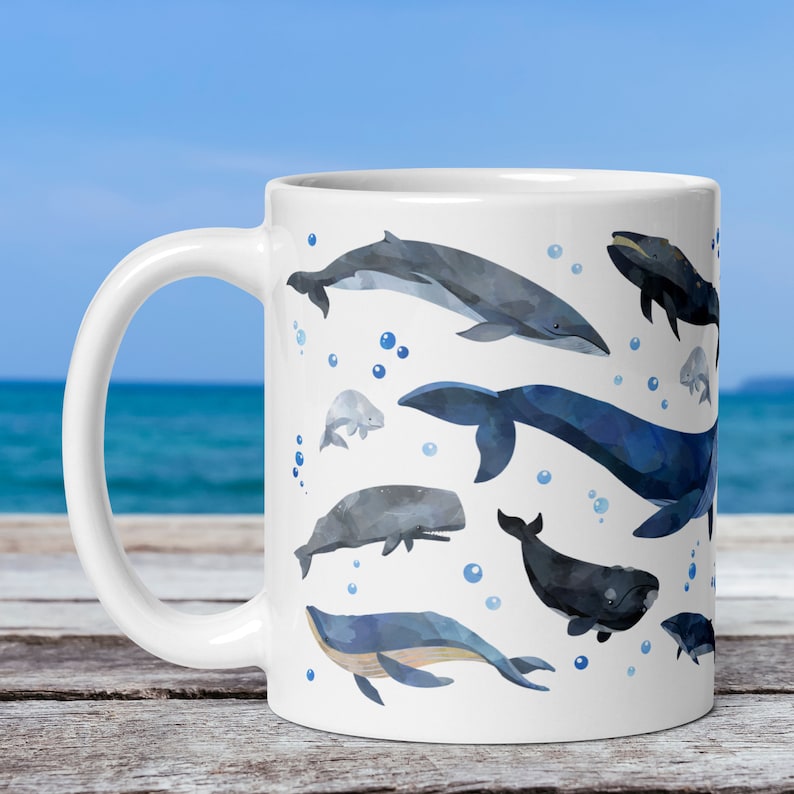 11-ounce ceramic mug with a watercolour whale print rests on a table with the ocean behind it. The mug's handle is on the left.