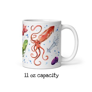 May include: White ceramic mug featuring watercolor illustrations of various squid species in shades of red, orange, green, and purple, with blue bubbles. The mug has an 11 oz capacity.