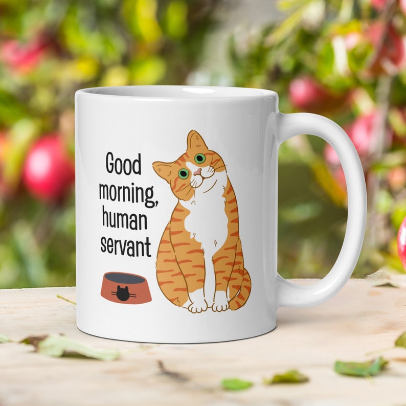A white ceramic mug depicting an orange tabby cat waiting expectantly by its empty food bowl, with the text "Good morning, human servant" printed above the bowl.