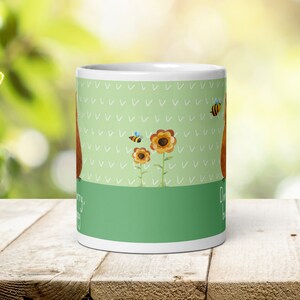 Op de afbeelding: Witte keramische mok met een groene achtergrond en een bruine cartoonillustratie van een beer met zonnebloemen en bijen. De tekst op de mok luidt "Maak je geen zorgen, wees gelukkig."