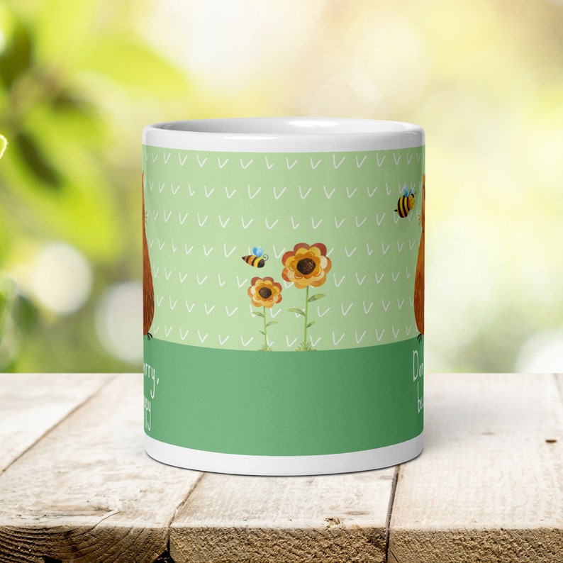 Op de afbeelding: Witte keramische mok met een groene achtergrond en een bruine cartoonillustratie van een beer met zonnebloemen en bijen. De tekst op de mok luidt "Maak je geen zorgen, wees gelukkig."