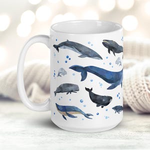 May include: White ceramic mug with a blue and gray watercolor design of various whales and dolphins. The mug has a large handle and is decorated with a repeating pattern of the sea creatures.