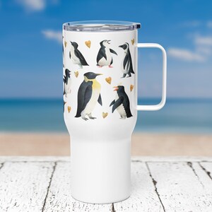 Peut inclure: Mug de voyage blanc avec une poignée noire et un motif de pingouins et de cœurs. Les pingouins sont noirs et blancs avec des becs orange. Les cœurs sont dorés.