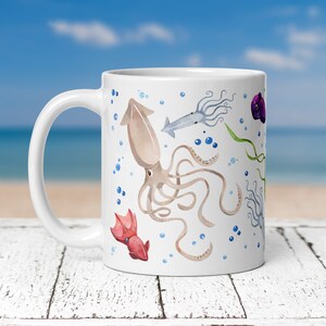 May include: White ceramic mug featuring a watercolor design of various squid and marine life. The design includes a large brown squid, smaller squid, bubbles, and seaweed. The mug has a curved handle and is set against a blurred beach background.