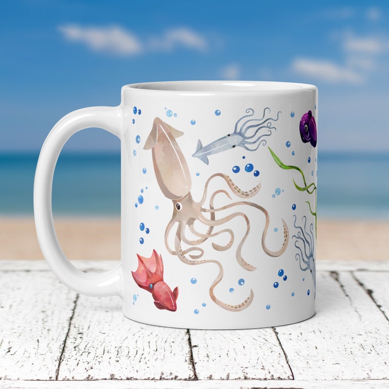 May include: White ceramic mug featuring a watercolor design of various squid and marine life. The design includes a large brown squid, smaller squid, bubbles, and seaweed. The mug has a curved handle and is set against a blurred beach background.