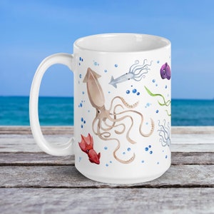 May include: White ceramic mug featuring watercolor illustrations of various squid species in shades of brown, red, and gray, along with blue bubbles and green seaweed. The mug has a large handle and a smooth, glossy finish.