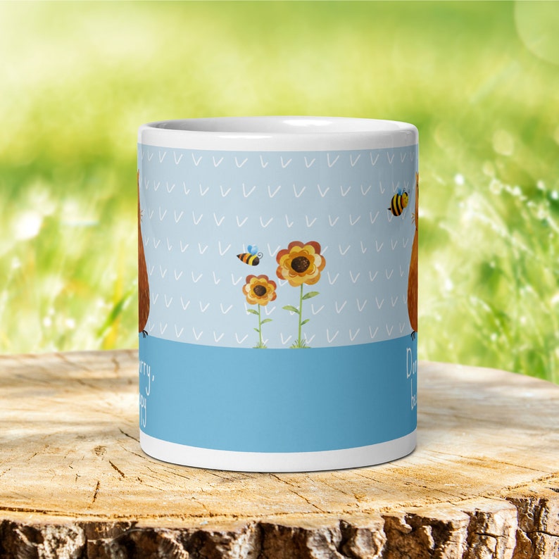 Op de afbeelding: Witte keramische mok met een blauw-wit patroon. De mok heeft een cartoonillustratie van een bruine beer met zonnebloemen en bijen. De tekst "Maak je geen zorgen, wees gelukkig" staat op de mok gedrukt.