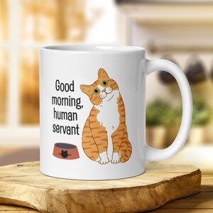 A white ceramic mug depicting an orange tabby cat waiting expectantly by its empty food bowl, with the text "Good morning, human servant" printed above the bowl.