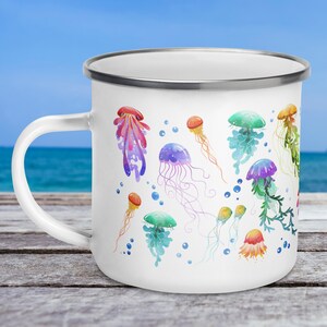 A 12-ounce enamel mug rests on an outdoor table with the ocean behind it. The mug has a silver rim and its handle is on the left. Printed on the white mug are colourful watercolour illustrations of jellyfish in pink, green, orange, purple and blue.