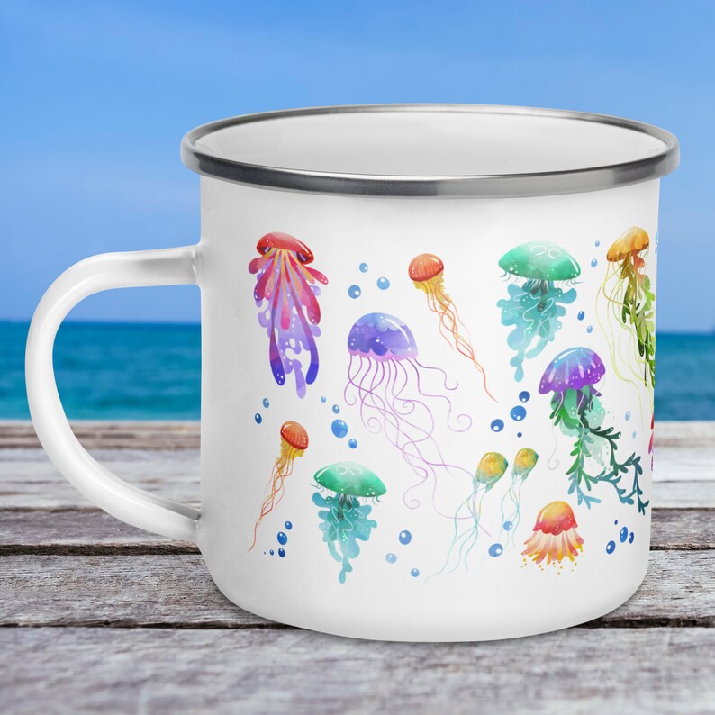 A 12-ounce enamel mug rests on an outdoor table with the ocean behind it. The mug has a silver rim and its handle is on the left. Printed on the white mug are colourful watercolour illustrations of jellyfish in pink, green, orange, purple and blue.