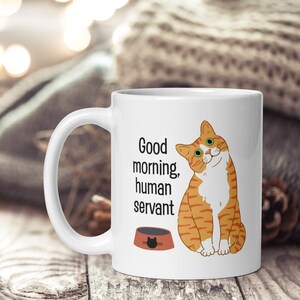 A white ceramic mug depicting an orange tabby cat waiting expectantly by its empty food bowl, with the text "Good morning, human servant" printed above the bowl.