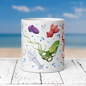 May include: White ceramic mug featuring watercolor illustrations of various sea creatures, including squid and octopus in shades of purple, red, green, and beige. Blue bubbles are scattered around the sea creatures. The mug is cylindrical and appears to be 11 ounces.