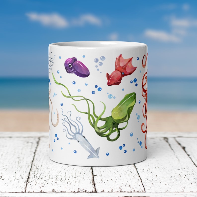 May include: White ceramic mug featuring watercolor illustrations of various sea creatures, including squid and octopus in shades of purple, red, green, and beige. Blue bubbles are scattered around the sea creatures. The mug is cylindrical and appears to be 11 ounces.