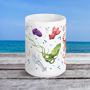 May include: White ceramic mug featuring colorful watercolor illustrations of various squid species, including purple, red, green, and gray varieties. The design includes bubbles and seaweed elements, set against a white background.
